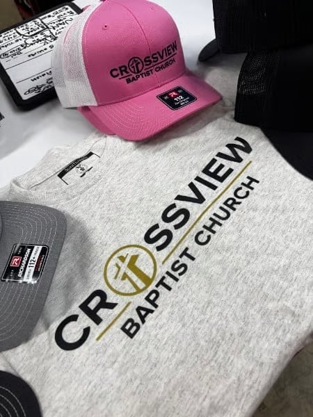 Pink and white Crossview Baptist Church branded cap and gray t-shirt with cross logo displayed on white surface