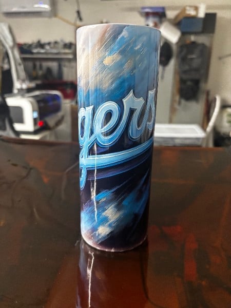 A cylindrical tumbler with blue and white design featuring what appears to be a sports team logo, sitting on a dark wooden surface in a workshop setting