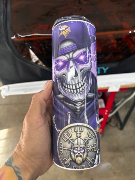 Hand holding a tall purple energy drink can featuring a skull design with purple bow and Viking medallion artwork