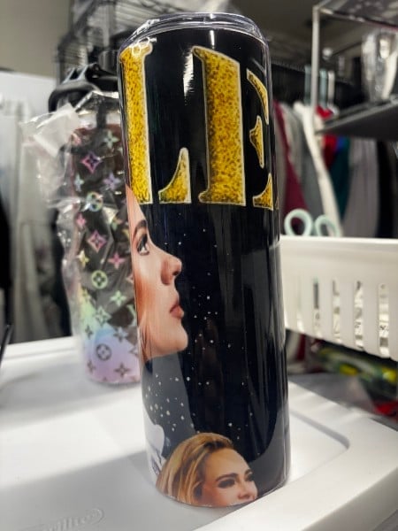 Black tumbler with gold lettering and portrait images of women displayed on white shelf with cluttered background