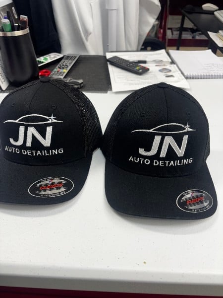 Two black baseball caps with JN AUTO DETAILING logo displayed on a white desk in an office setting