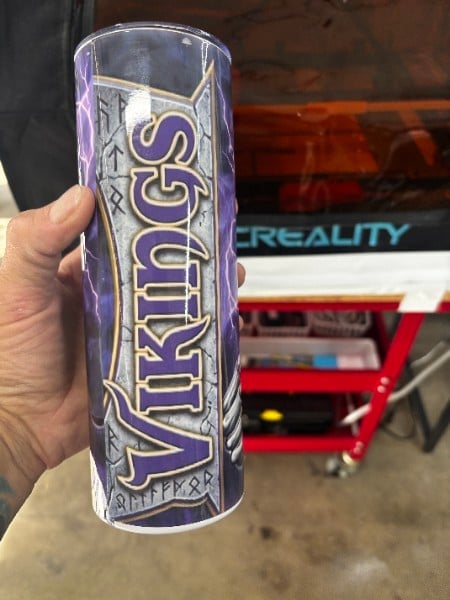 Hand holding a tall purple and silver Vikings tumbler in a garage setting with red toolbox visible in background