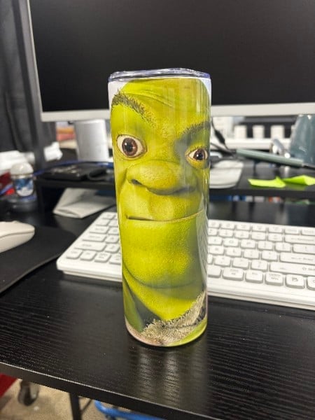 Novelty yellow drinking glass featuring a smiling shrek-like character face, sitting on a desk with computer equipment in background