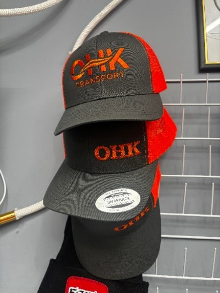 Stack of four OHK Transport branded baseball caps in red and black colors displayed on a mannequin head