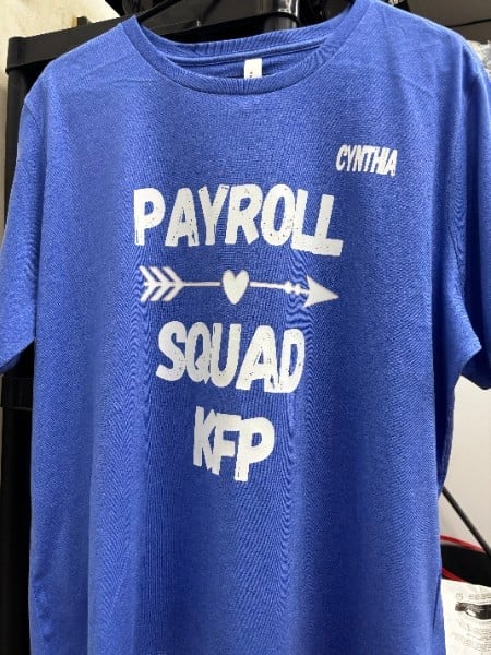 Blue t-shirt with white text reading Payroll Squad with arrows and heart design, personalized with names Cinthia and KFP