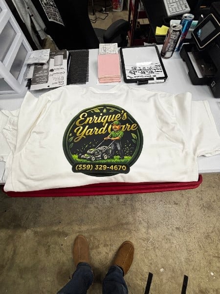 White t-shirt with Enriques Hardware logo and phone number displayed on table at printing shop